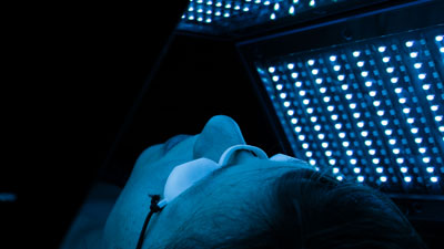 Une femme en pleine séance de photobiomodulation LED sur le visage et sous une lumière bleue.