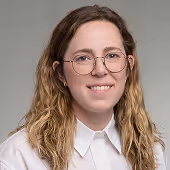Smiling woman with long wavy hair and glasses wearing a white collared shirt against a gray background.