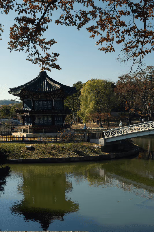 Traditional pavilion surrounded by trees and water, with a bridge and reflections on a lake on a clear day.