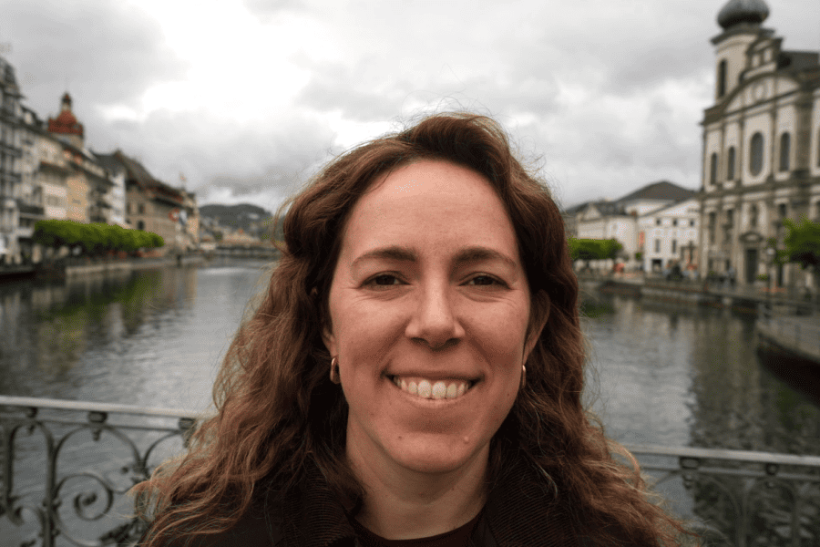 Woman smiling on a bridge over a river with historic buildings and cloudy sky in the background.