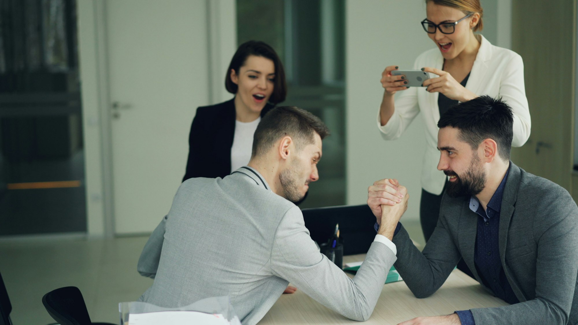 Colleagues arm wrestling while others watch and film — team building fun in the office. Photo by Vitaly Gariev on Unsplash