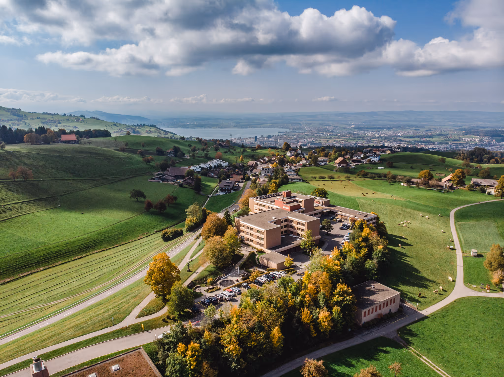 Luftaufnahme eines Gebäudekomplexes inmitten grüner Hügel mit verstreuten Häusern und einer Stadt am Horizont unter bewölktem Himmel.