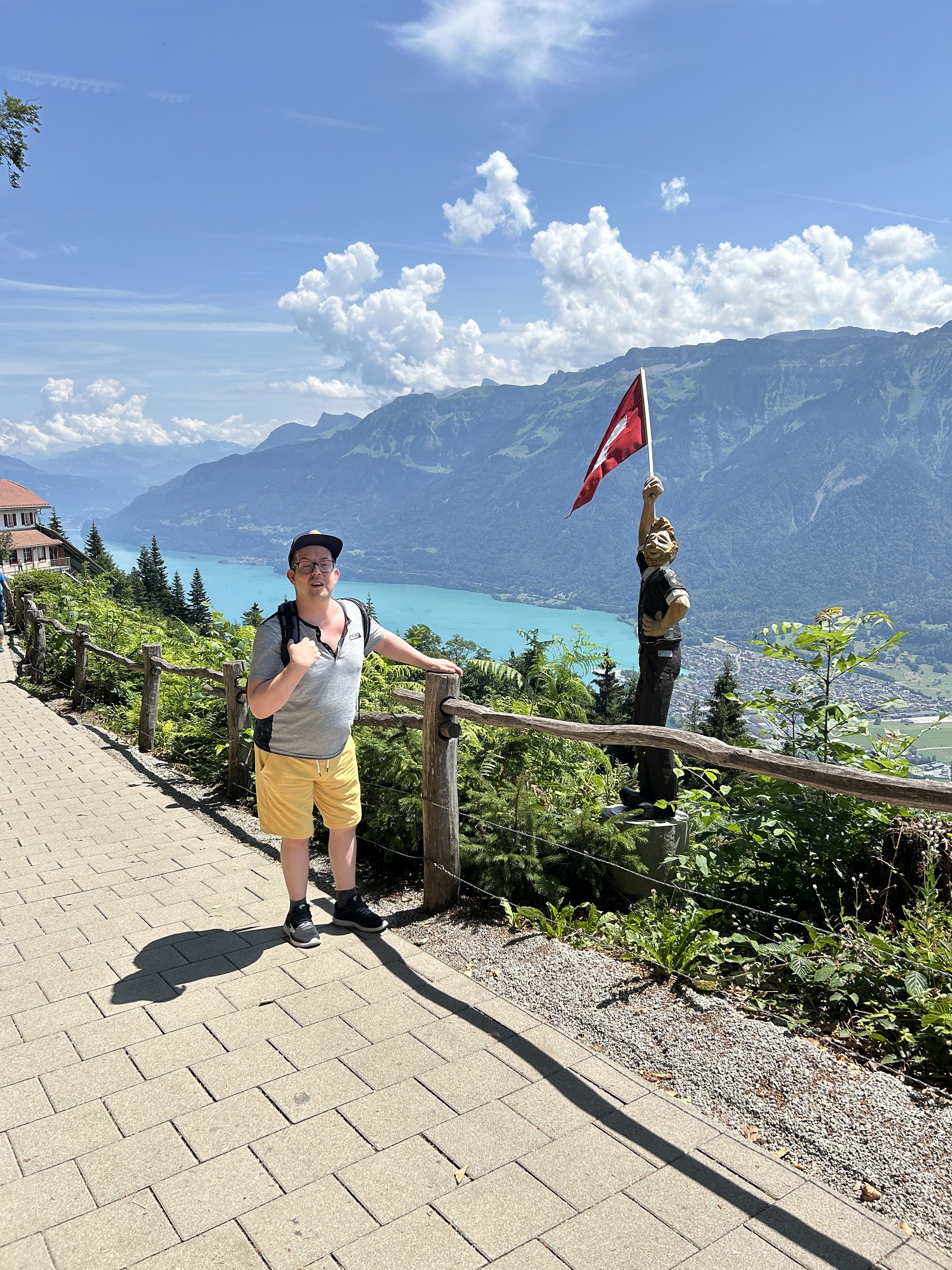 Mann mit Rucksack vor dem Brienzersee, daneben eine Holzfigur mit Schweizer Fahne