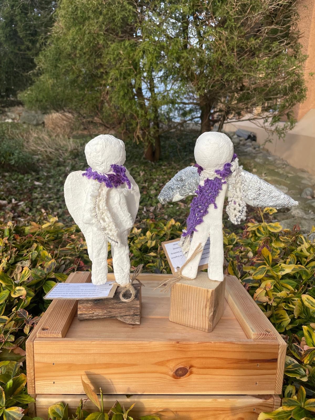 Zwei weisse Engelsfiguren mit violetten Schals auf einem Holzsockel im Garten