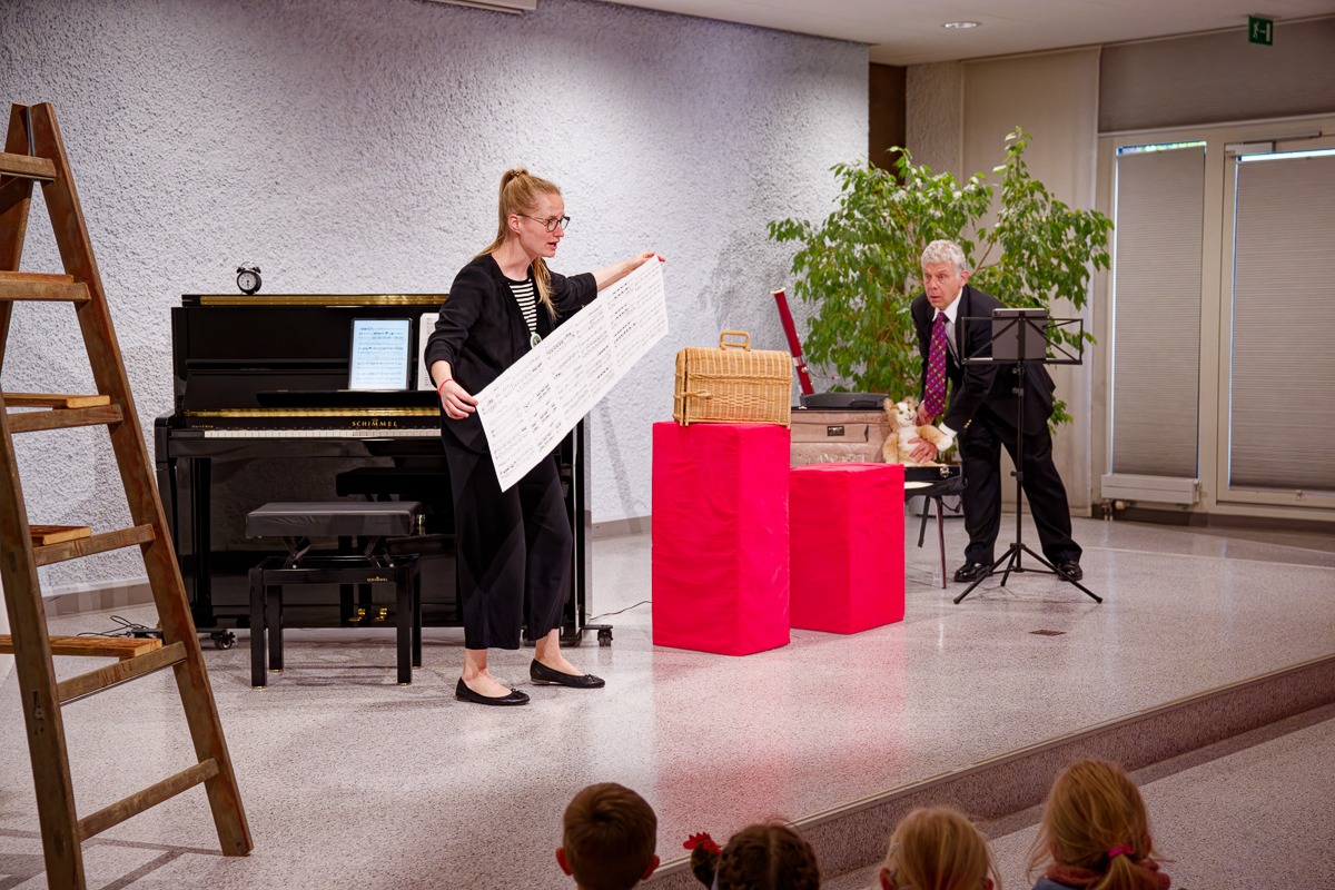 Konzert auf einer Bühne mit Klavier: Musikerin zeigt ein grosses Notenblatt, Kinder schauen zu