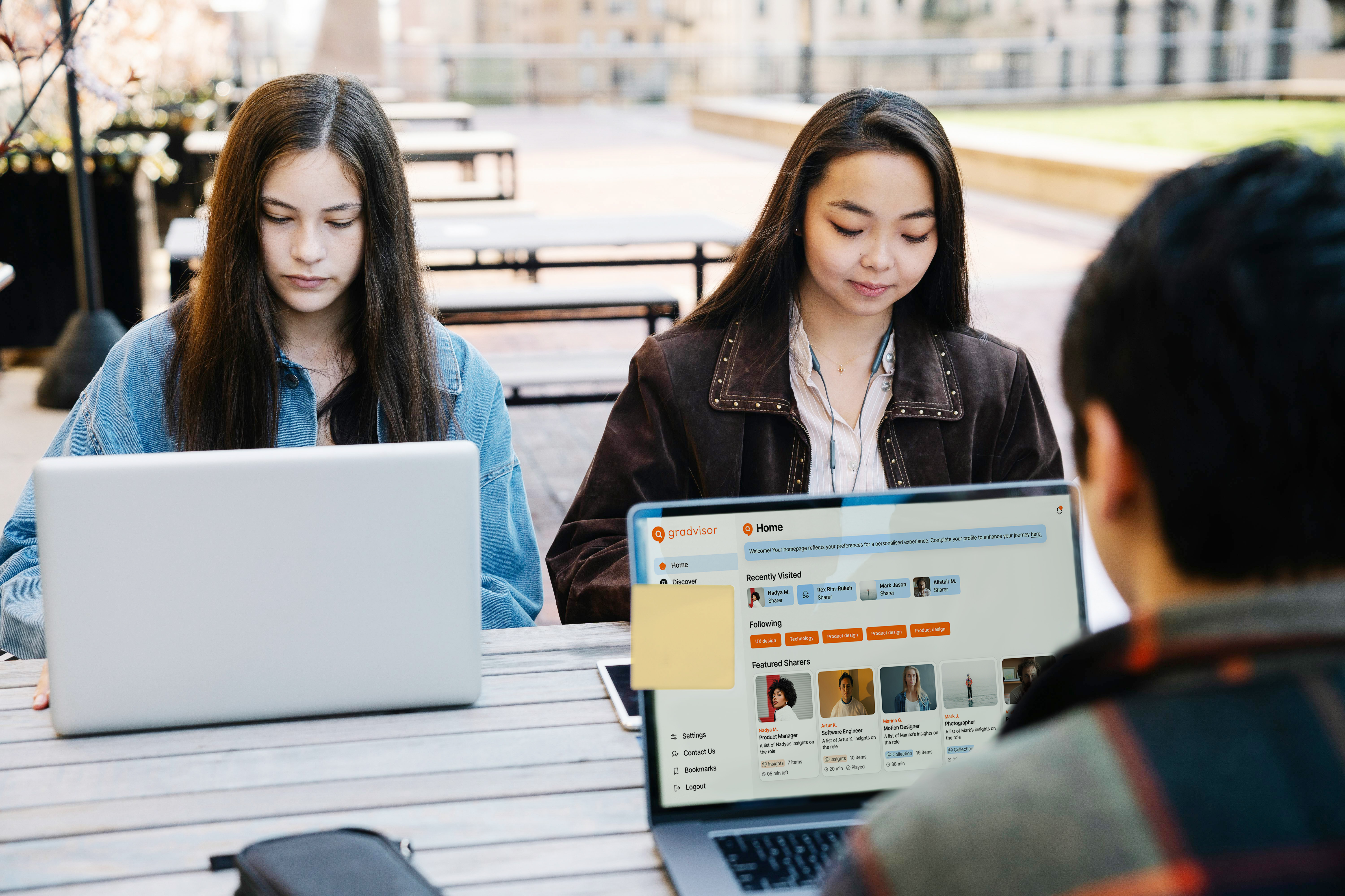 Three young adults working on laptops at an outdoor table with one laptop screen displaying a social networking site called Gradvisor.
