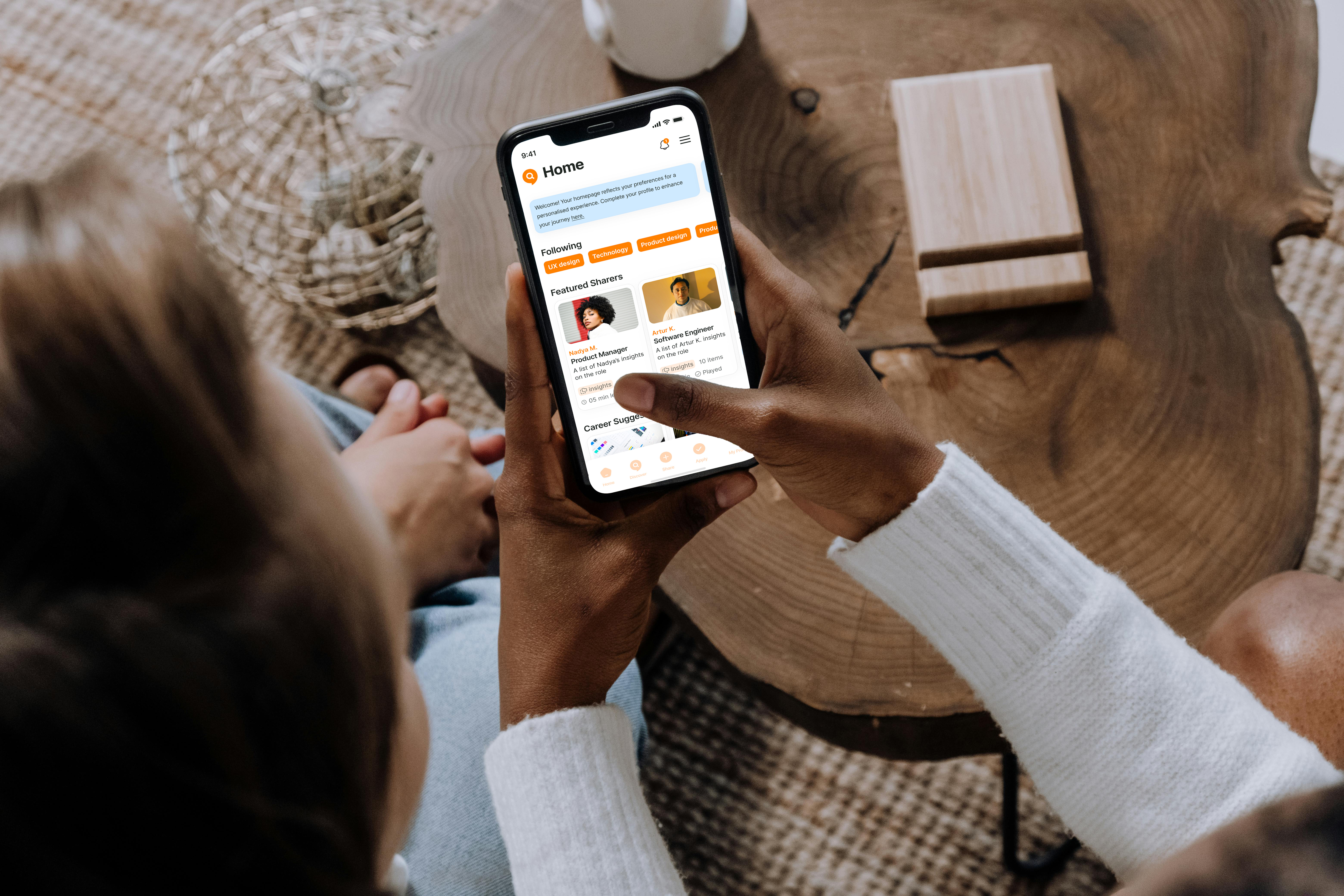 Person holding a smartphone showing a home feed with featured sharers and career suggestions, sitting by a wooden table.