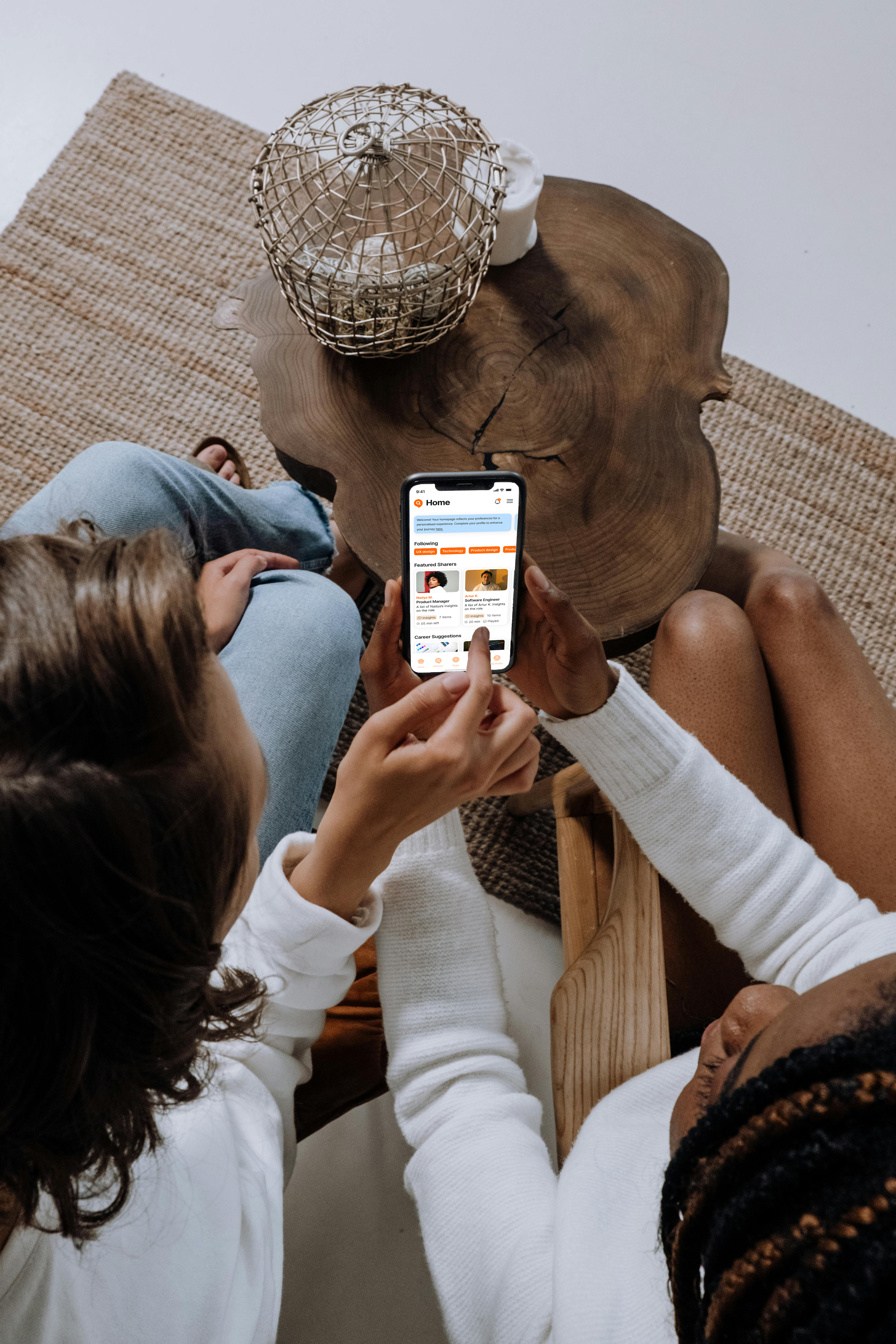 Two people sitting near a wooden table, looking at a smartphone screen showing a social media app interface.