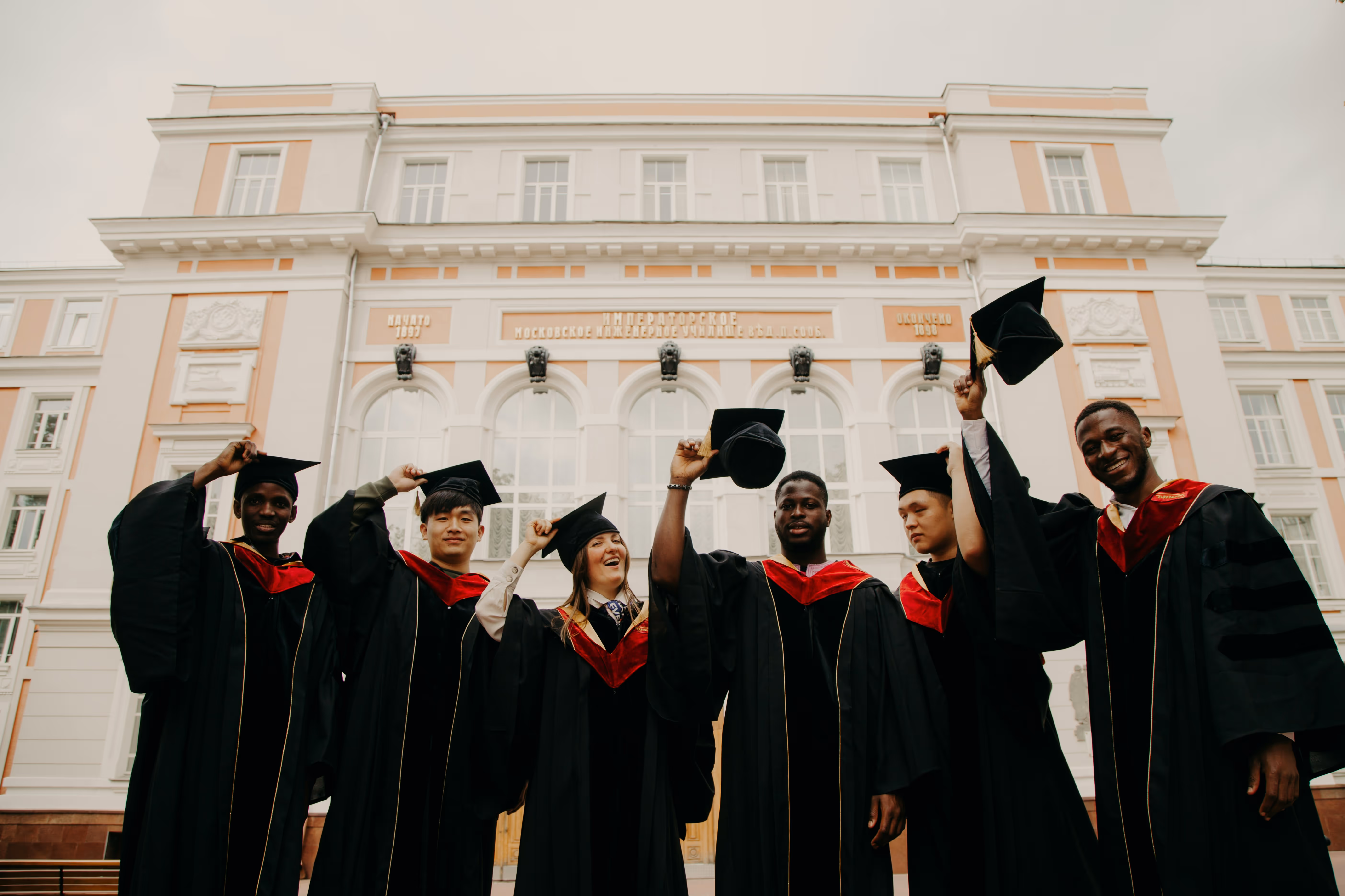Group of six diverse graduates in black caps and gowns holding their caps up in front of a large classical building.