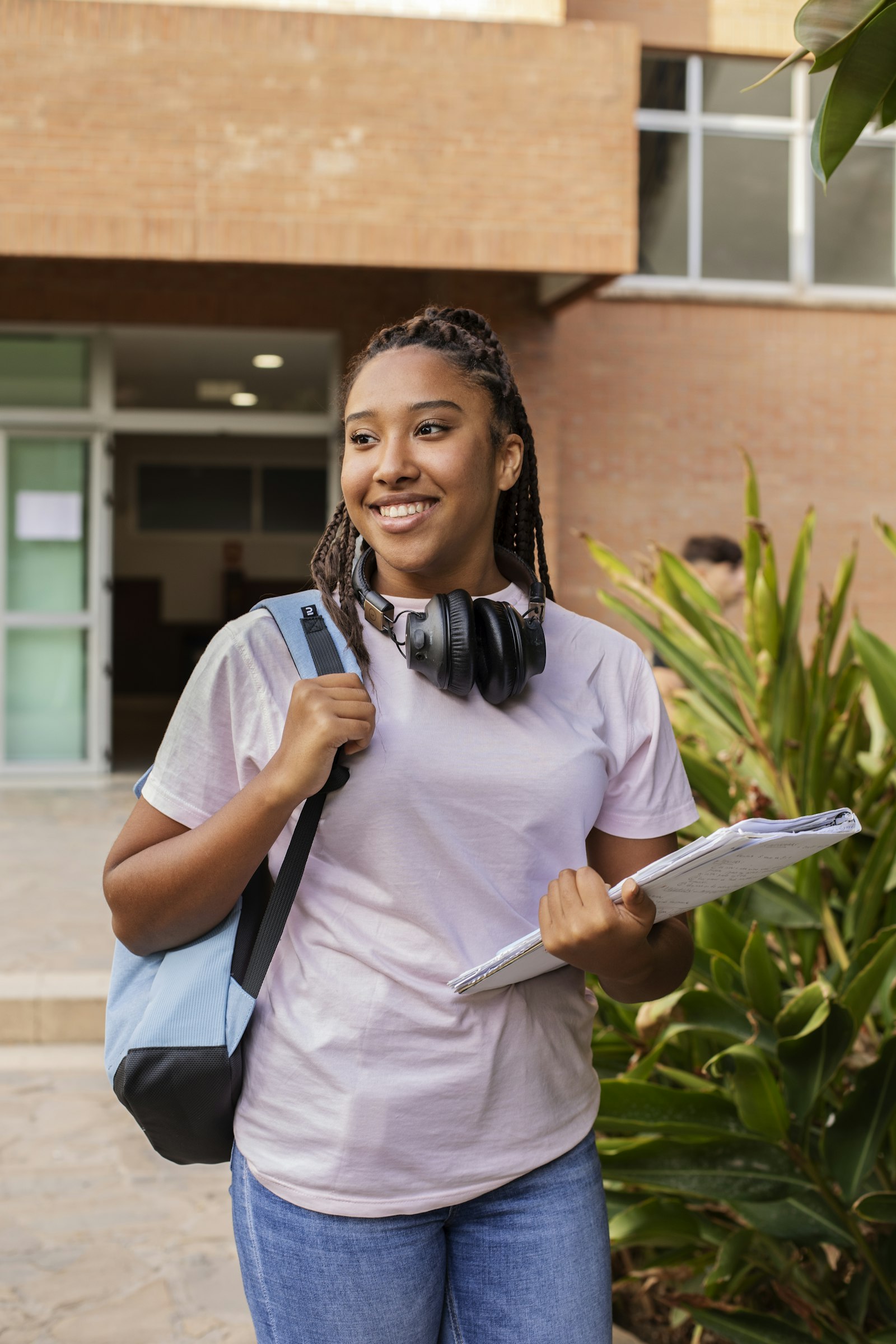 Smiling student on a university campus with headphones around her neck, a backpack and notes