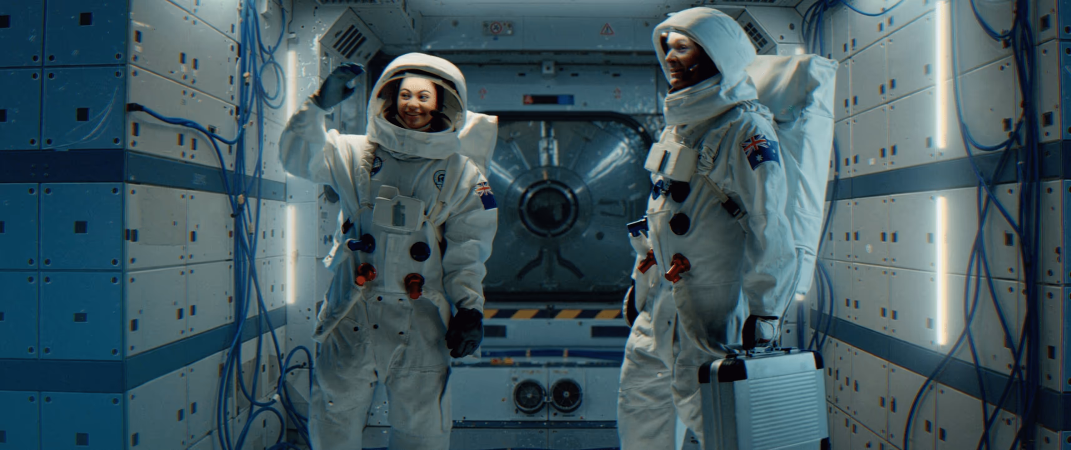 Two astronauts - one female Kiwi and one male Aussie - wave good bye while standing in a spaceship airlock.