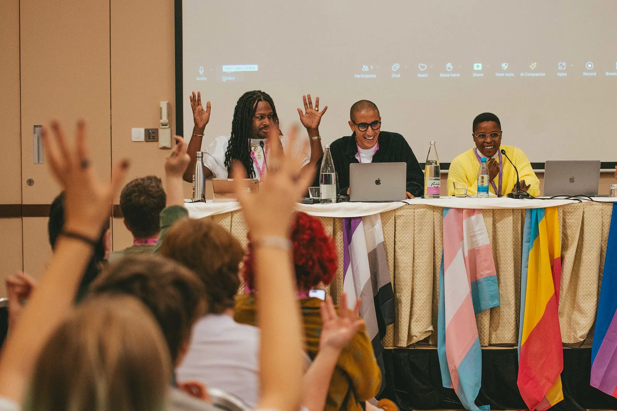 Photo of the Anti-Racism Panel at the IGLYO AMC Ljubljana 2024 where three laughing bipoc speakers are sitting at a table in front of audience members with their hands raised.