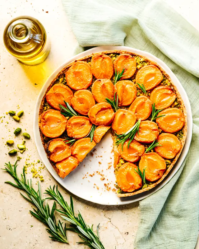 Stylisme culinaire d'une tarte à l'abricot et au romarin dans une ambiance chaleureuse avec une bouteille d'huile d'olive, des branches de romarin et des pistaches