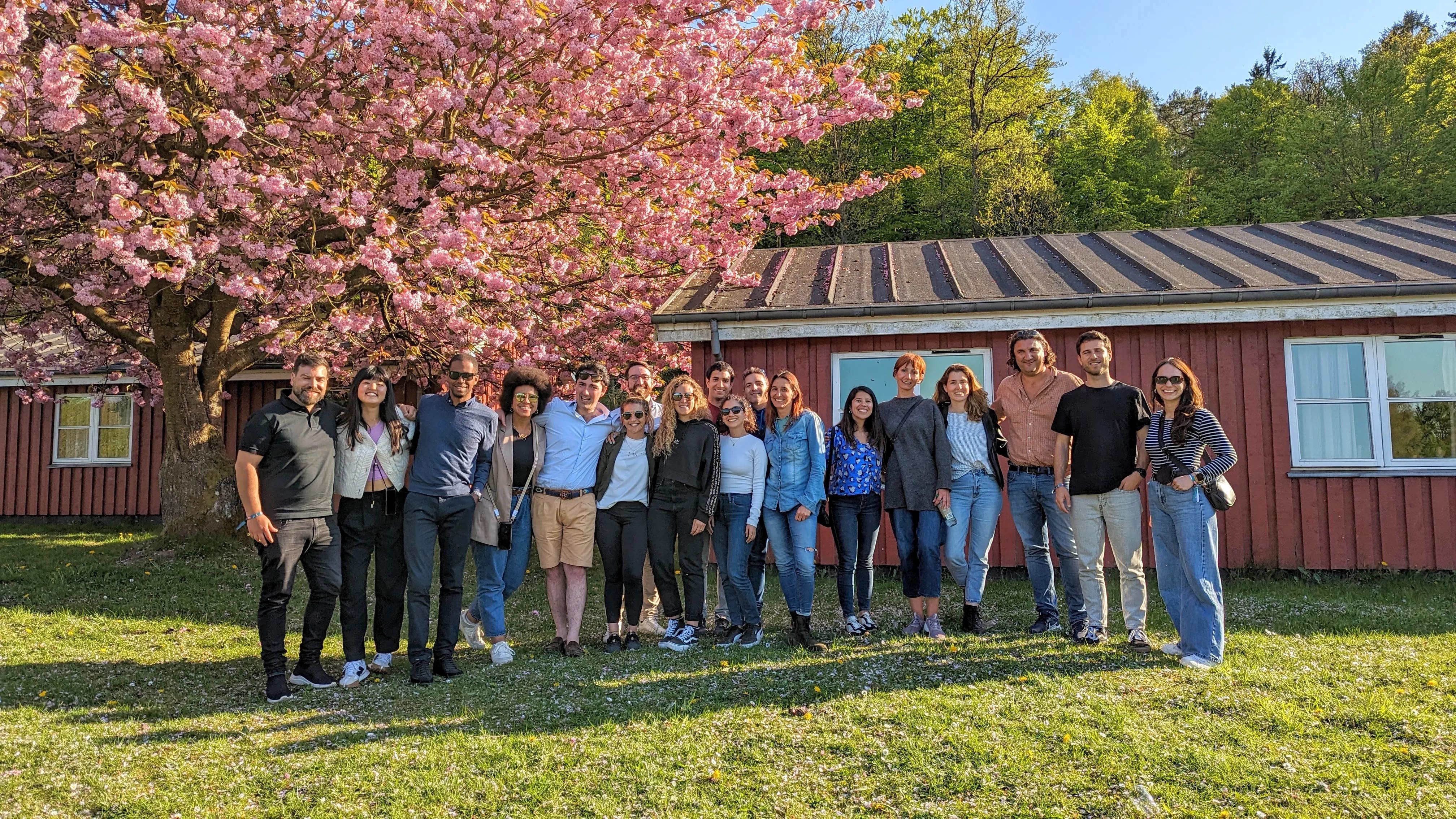 Grupo de dieciséis personas sonrientes posando juntas al aire libre frente a una casa de madera roja con ventanas blancas y un árbol con flores rosas en primavera.