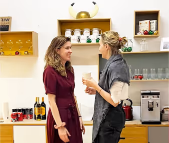 Dos mujeres conversando frente a una máquina de café en un ambiente interior moderno.