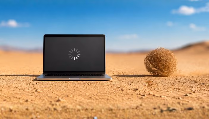 Portátil abierto en mitad de un desierto soleado, con un símbolo de carga en la pantalla y una bola de arbusto seco a un lado.