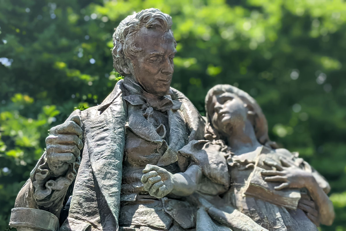 Bronze statue of a man holding a young girl who clutches a book against a background of green trees.