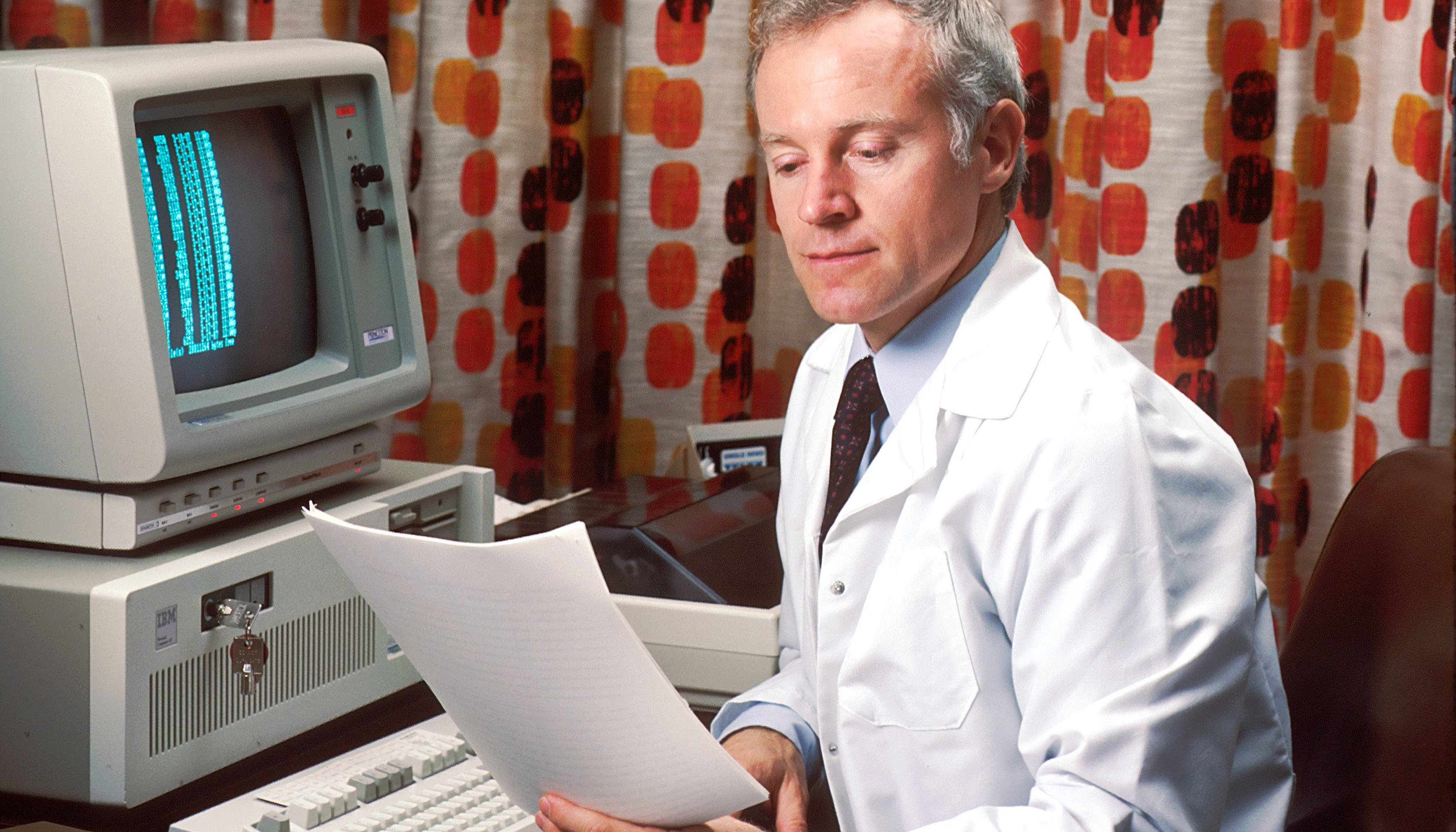 A man in a white coat reading a printout beside a 1980s computer terminal