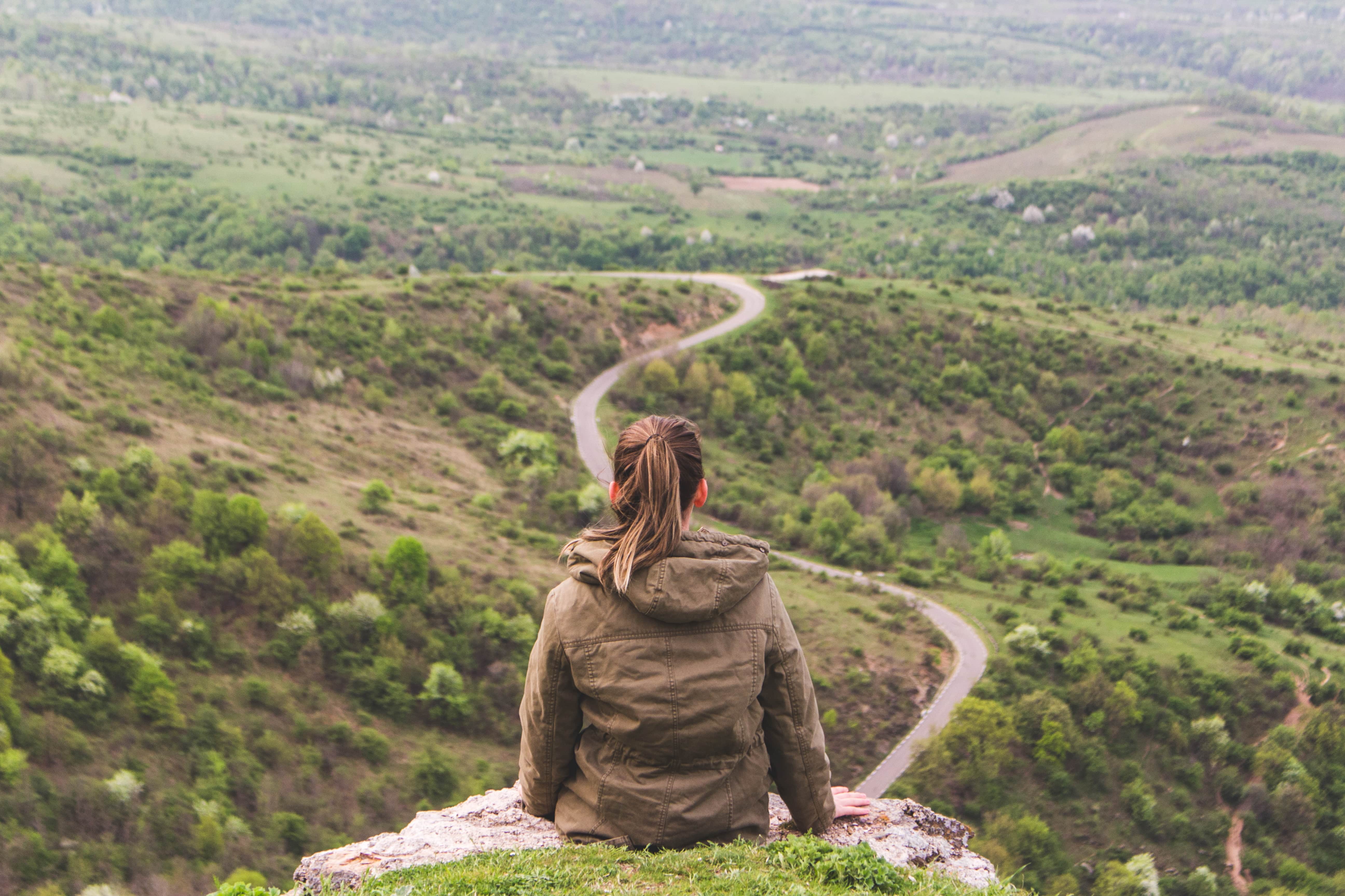 A person sitting on a hillside, looking out over a valley and a winding road