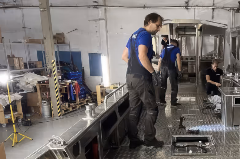 Three workers wearing blue shirts and black overalls assembling or inspecting a large metal structure inside an industrial workshop.