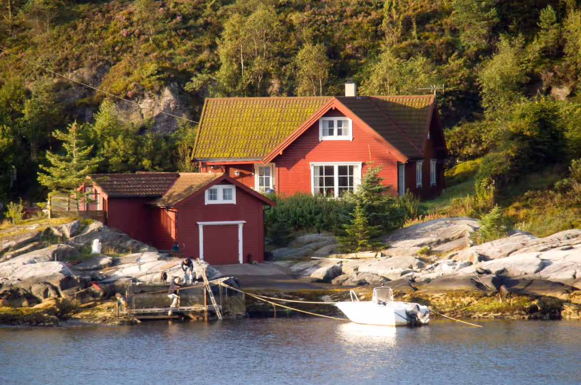 Red wooden cottage with a mossy roof by rocky shoreline, a small dock with people, and a white boat tied to shore.
