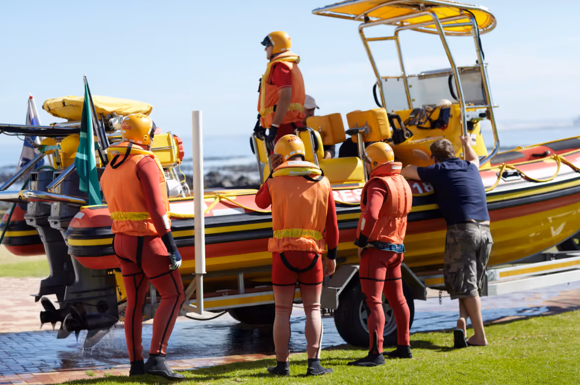 Four rescue workers in orange life vests and helmets preparing a yellow rescue boat by the shore.
