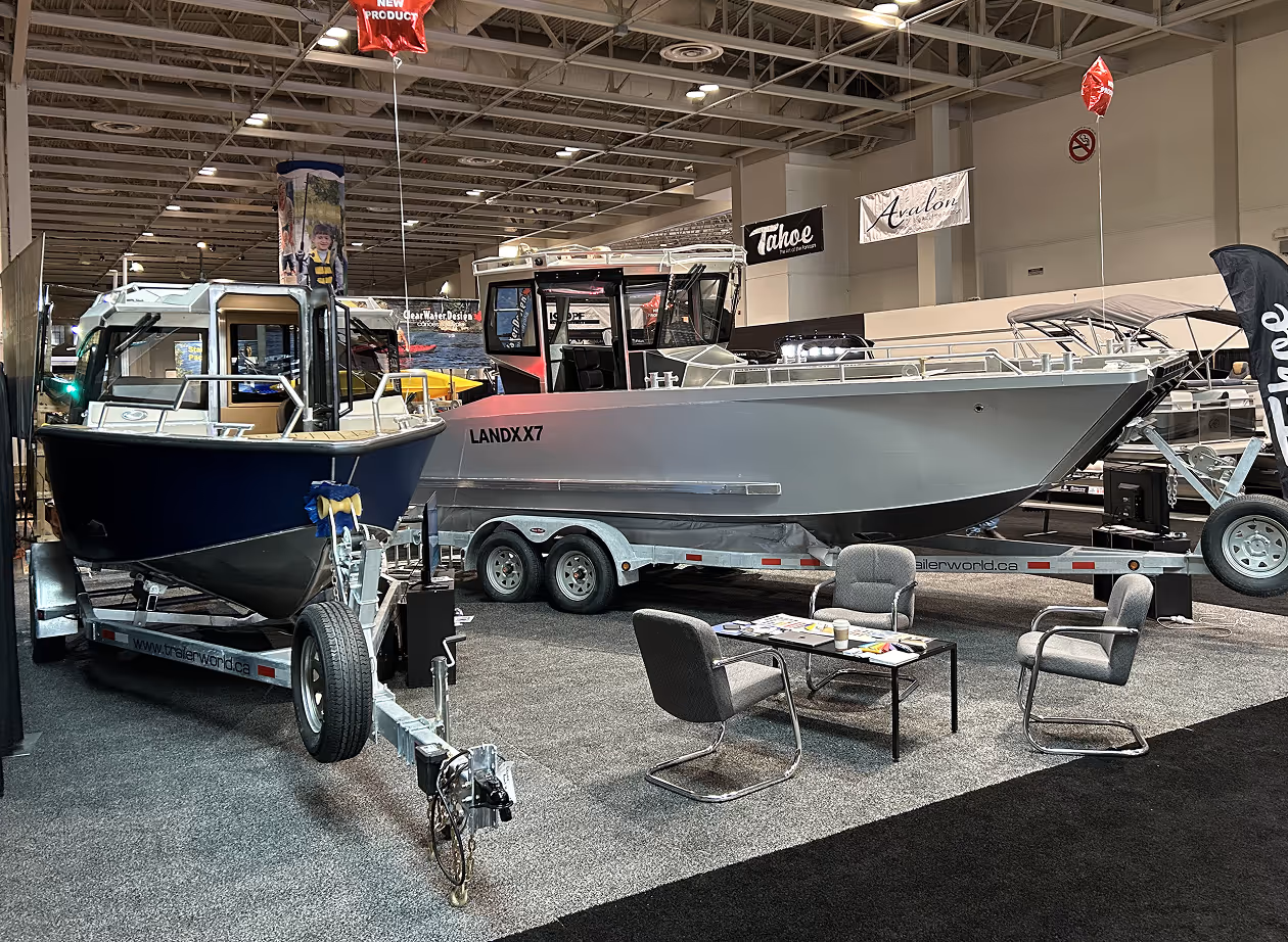 Two boats displayed on trailers at an indoor boat show with chairs and a table nearby.
