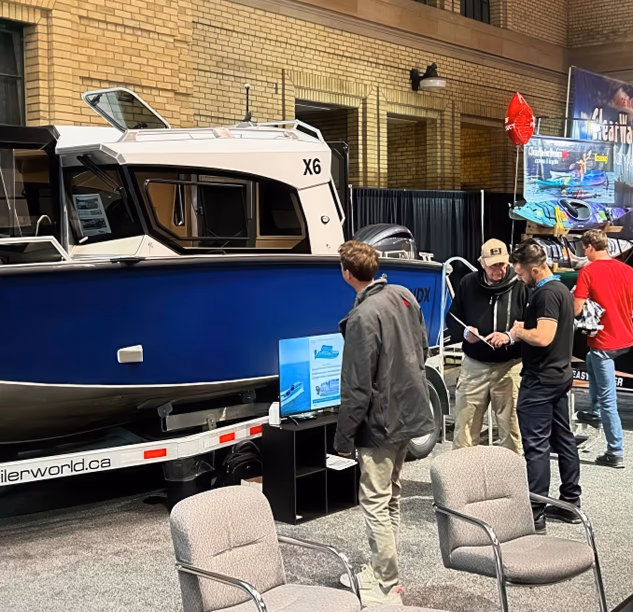 People examine a blue and white X6 motorboat displayed indoors at a trade show with chairs and informational screens nearby.