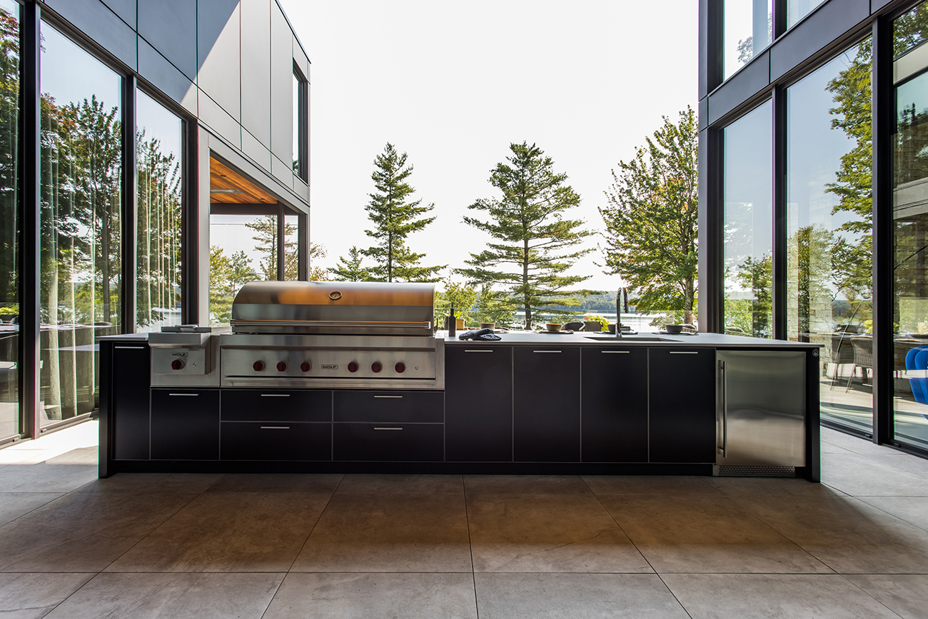 Modern outdoor kitchen with stainless steel grill, black cabinetry, and a lake view through large glass windows.