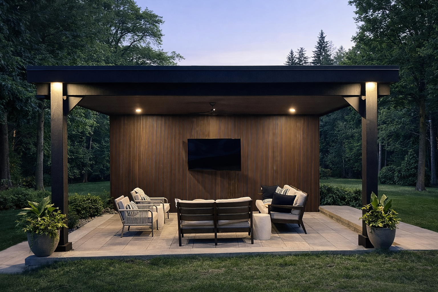 Modern outdoor patio with cushioned seating, a mounted flat-screen TV, ceiling fan, and ambient lighting under a covered pergola surrounded by greenery.
