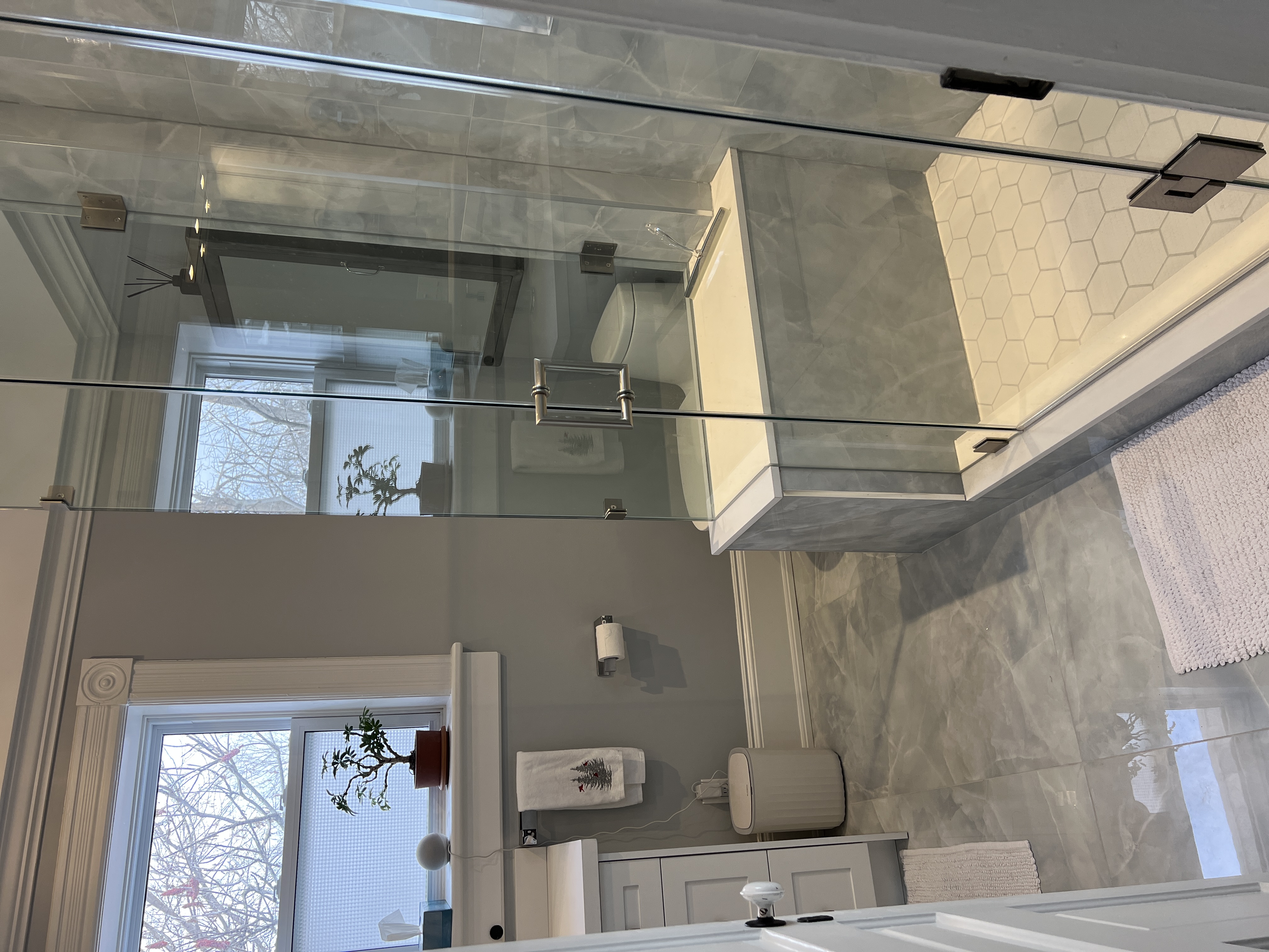 Modern bathroom with a glass shower enclosure, hexagonal floor tiles inside the shower, gray marble floor, a window with a potted plant, and white bath mats.