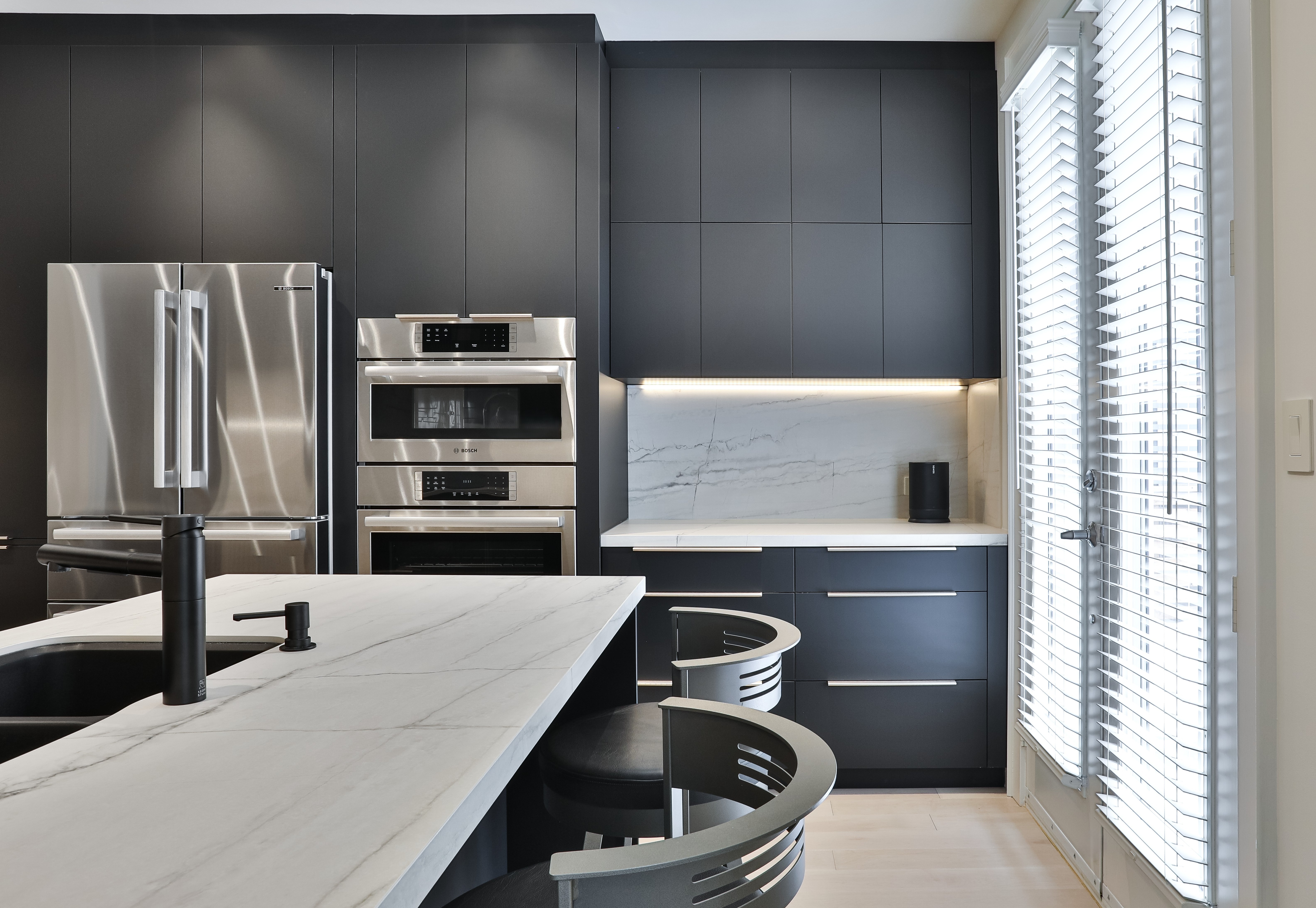 Modern kitchen with dark cabinetry, stainless steel appliances, marble countertops, black sink faucet, and large window blinds.