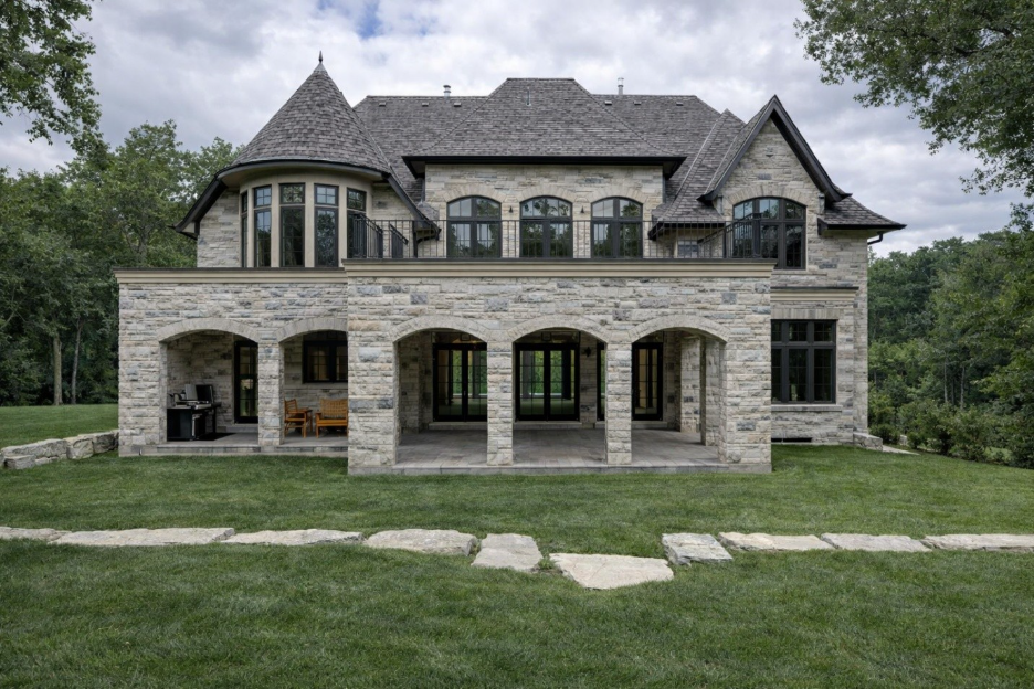 Stone two-story house with arched covered patio, turret, large windows, and a green lawn in front.