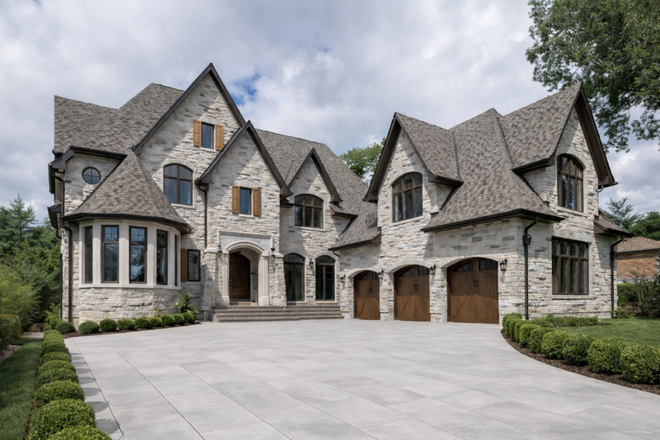Large two-story stone house with pitched roofs, arched windows, wooden garage doors, and a wide driveway.