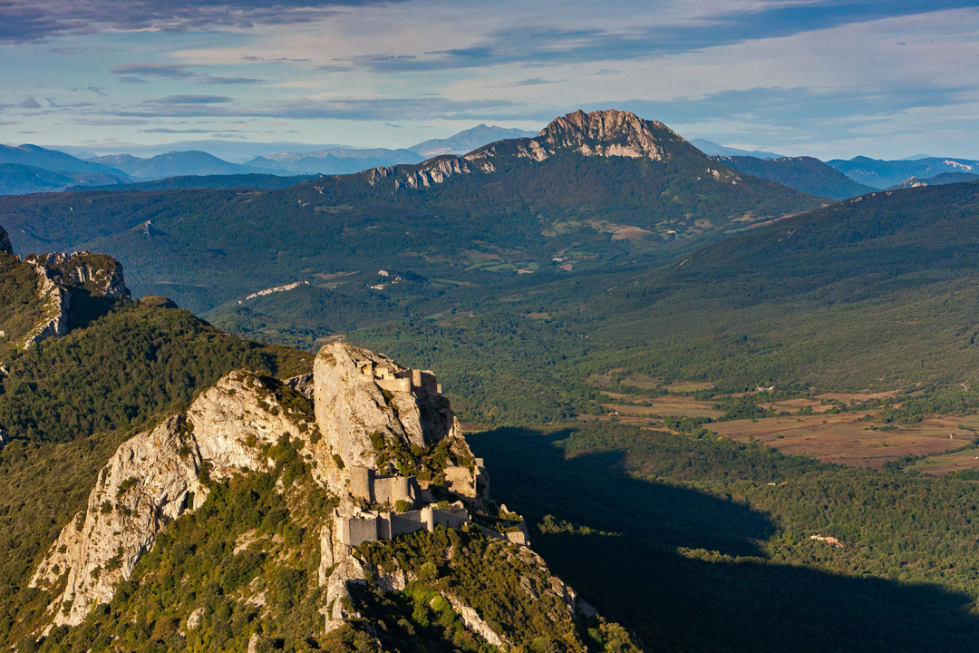 Aerial view of an ancient stone fortress perched on a rocky hill surrounded by forested mountains and valleys under a partly cloudy blue sky.