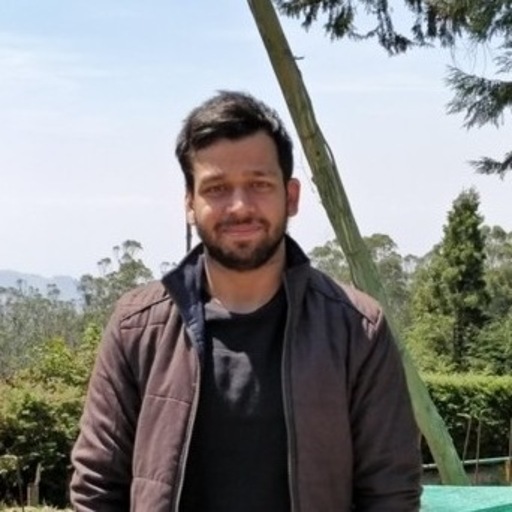 Man with short dark hair and beard wearing a brown jacket and black shirt outdoors with trees in the background.