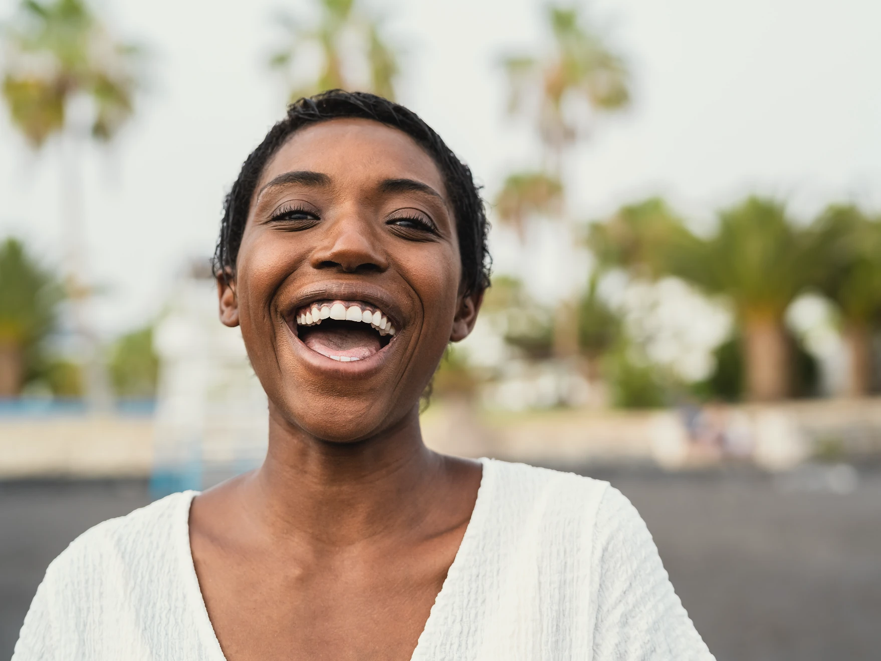 A woman smiling widely with palm trees blurred in the background.