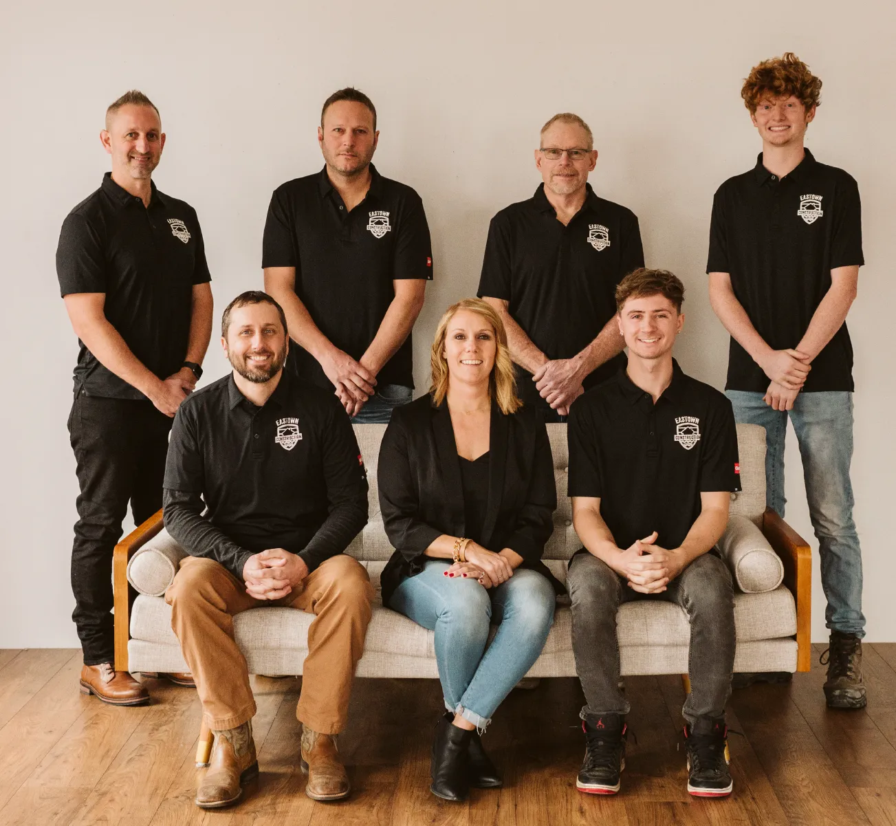 Group of seven people wearing Eastown Construction black shirts posing in a studio, three seated on a couch and four standing behind.