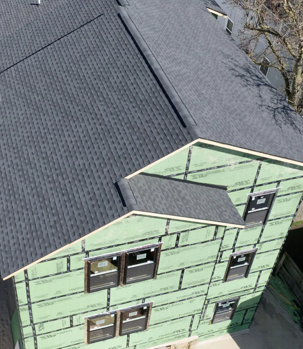 Aerial view of a partially constructed two-story house with green sheathing and new dark gray shingle roofing.