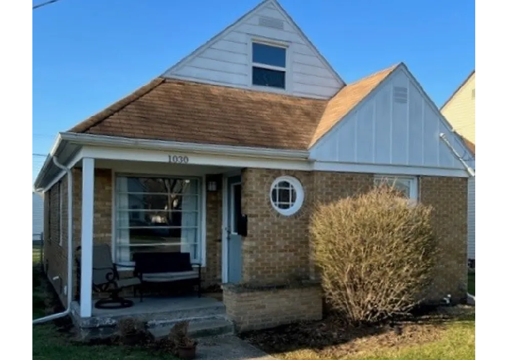 Small brick house with a worn brown roof, white trim, a front porch with chairs, and a large bush near the entrance.