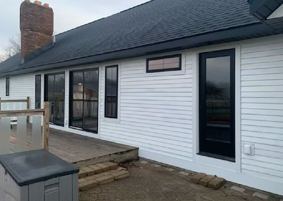 Side view of a house with white siding, a brick chimney, large windows, a black-framed door, and a wooden deck with stone steps.