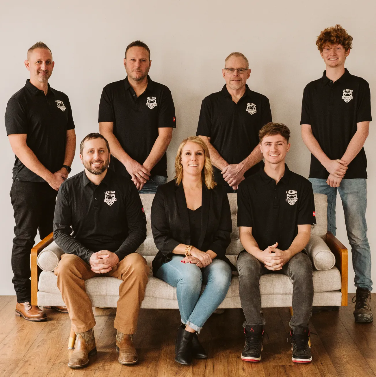 Eight people posing for a group photo, most wearing black Eastown Construction shirts, three seated on a beige couch and five standing behind it against a plain wall.