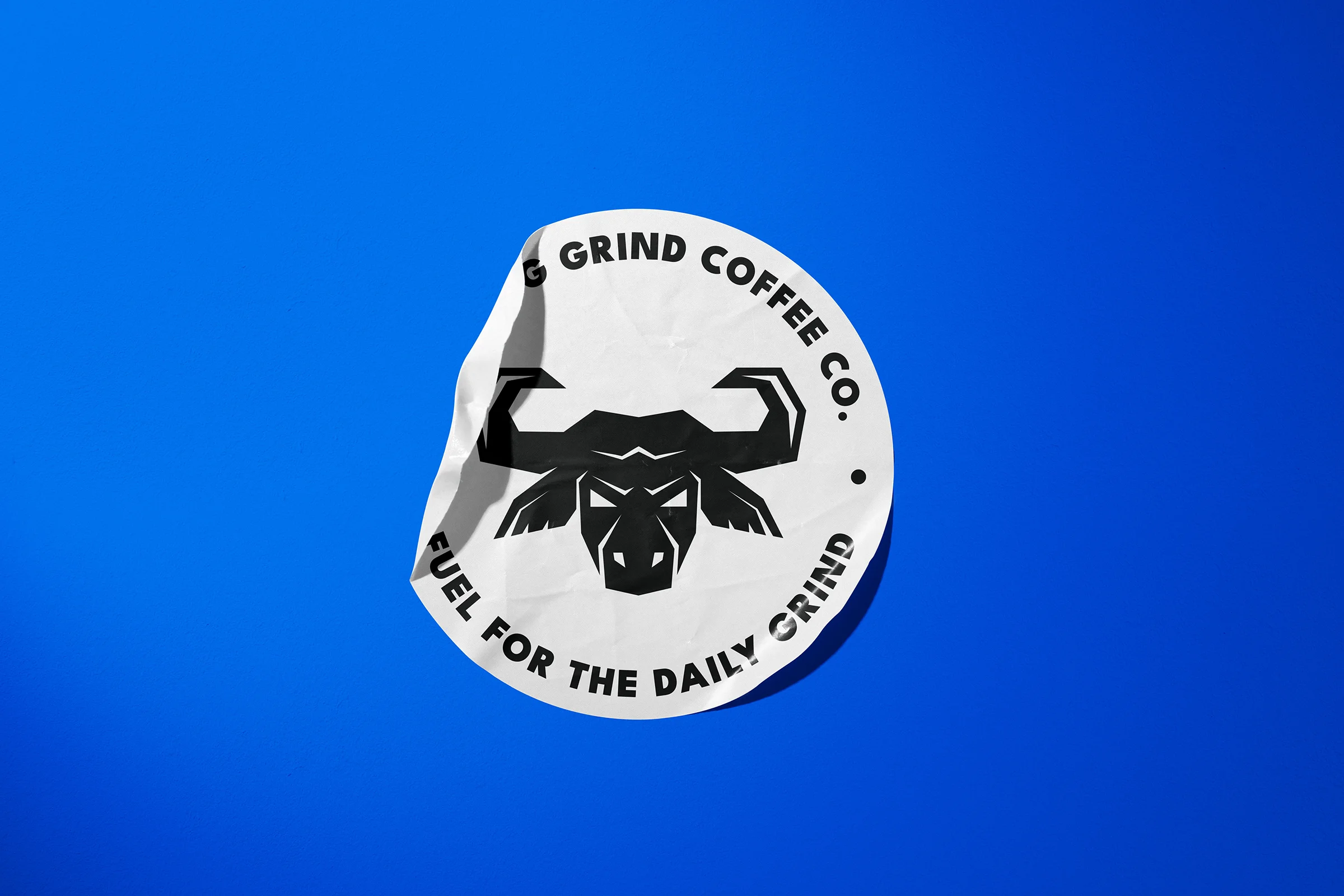A round, white paper sticker on a royal blue background. On the sticker is an angular geometric logo of a bull head. Around the edge of the sticker are the words Morning Grind Coffee Fuel For The Daily Grind.