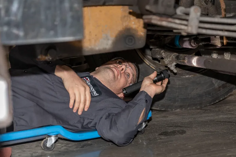 Suspension inspection being performed by a technician under a heavy-duty truck using a flashlight.