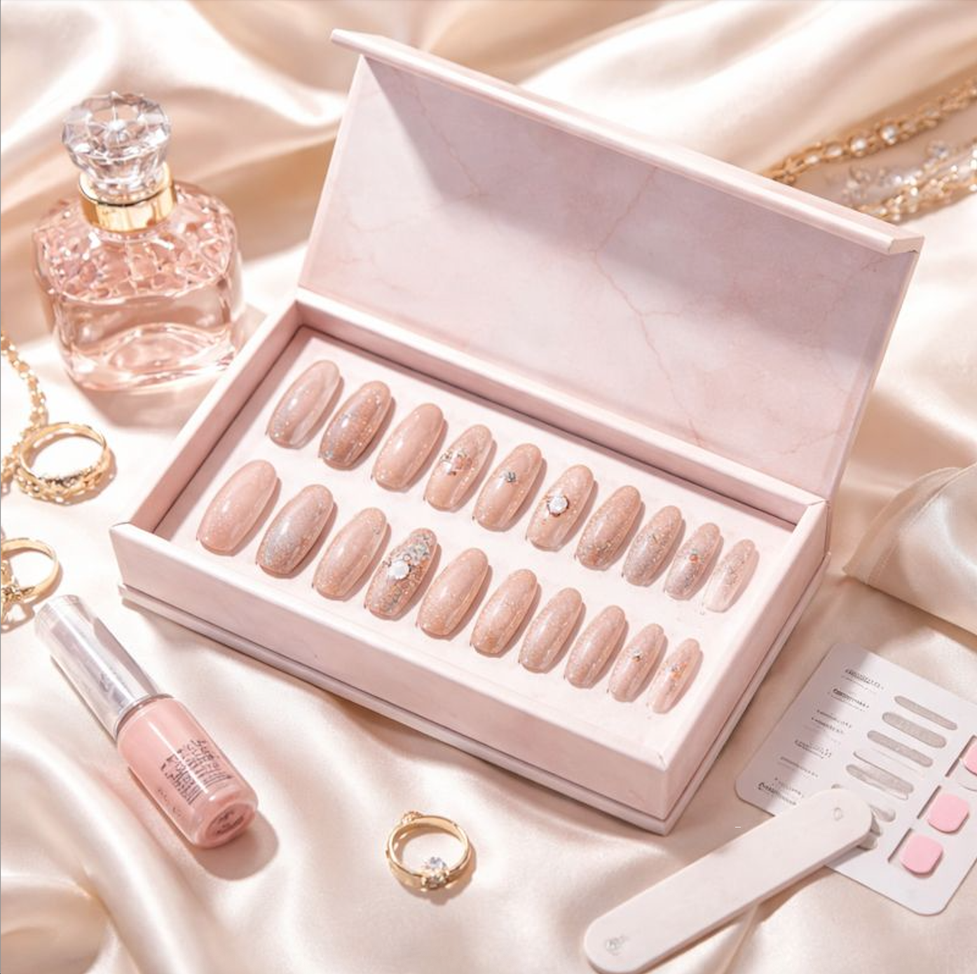 Elegant press-on nail set displayed in a premium presentation box, featuring glossy nude and glitter finishes with refined accessories in a soft, luxury flat-lay setting.
