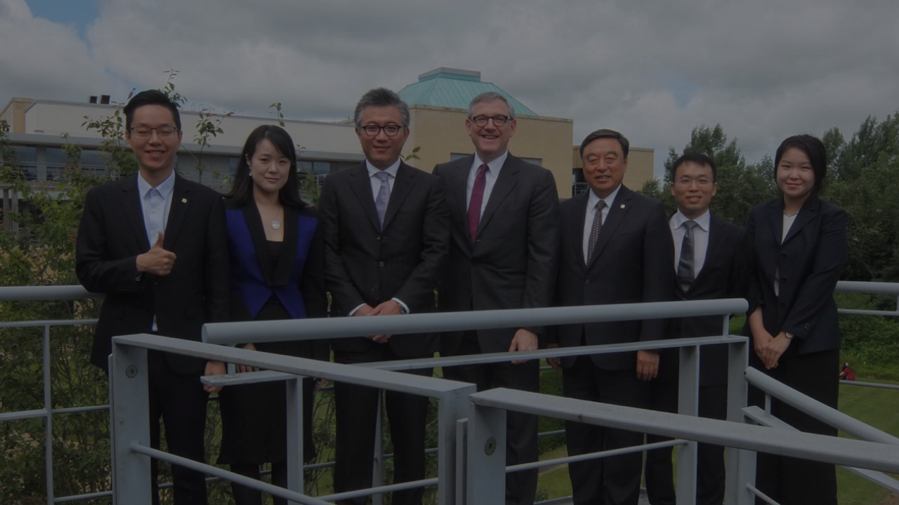Seven professionally dressed people standing outdoors on a balcony with greenery and buildings behind them.