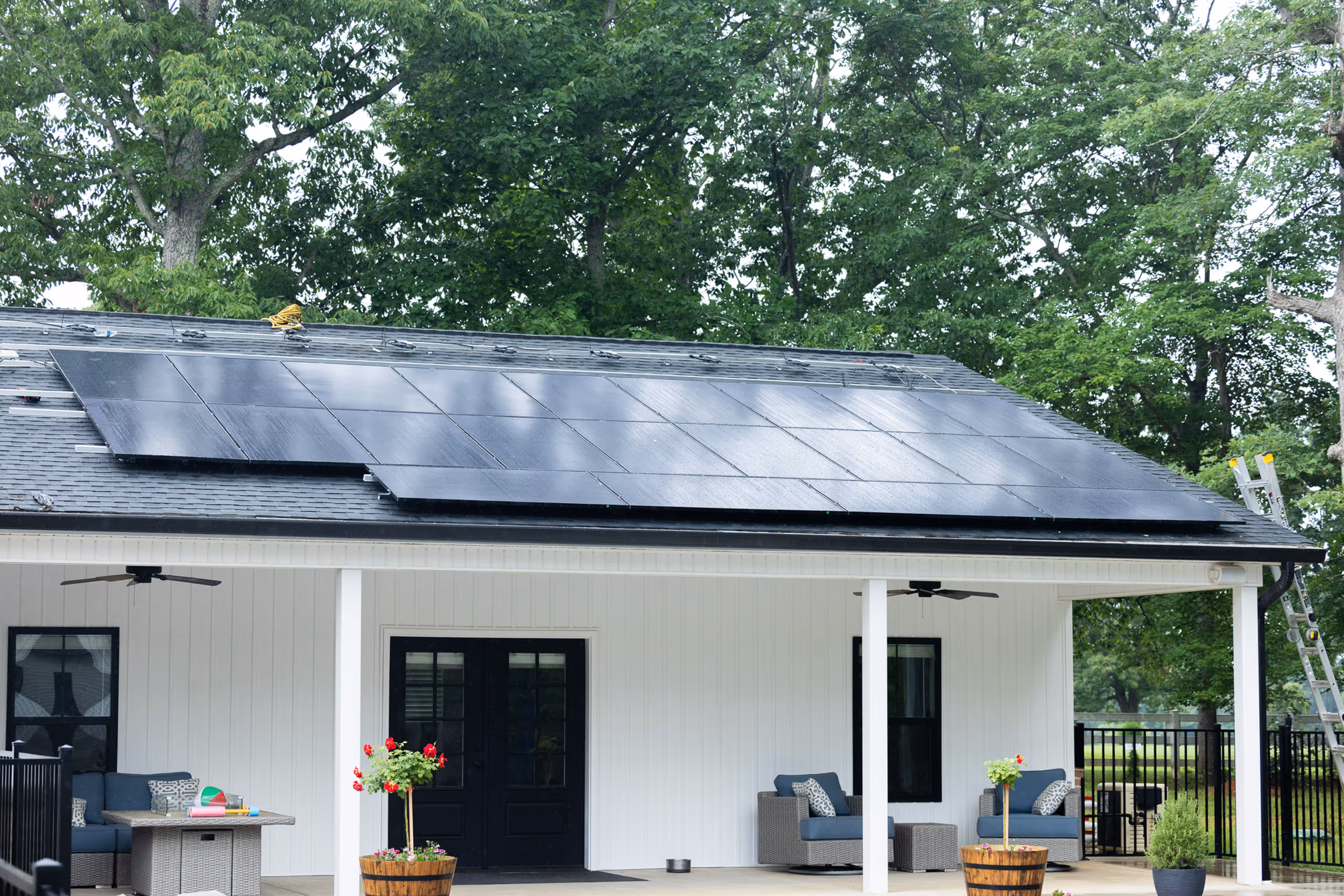 Black solar panels installed on the roof of a white house with a covered porch and outdoor seating.
