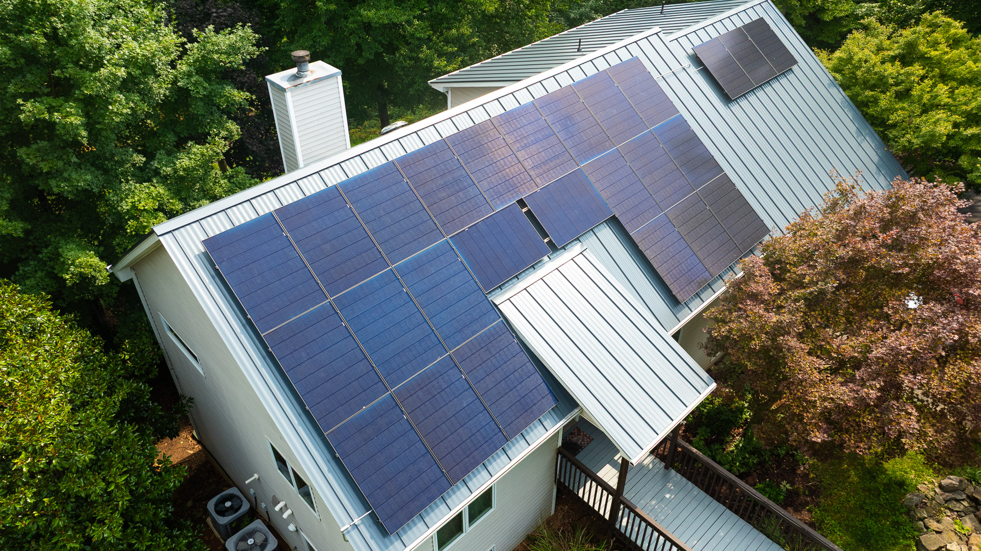 Black solar panels installed on the roof of a white house with a covered porch and outdoor seating.