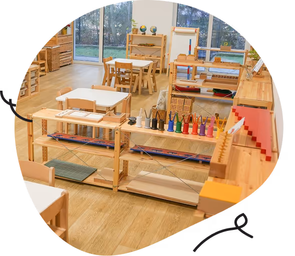 Przestronna sala przedszkolna z drewnianymi meblami, małymi stolikami i zestawem edukacyjnych materiałów Montessori.