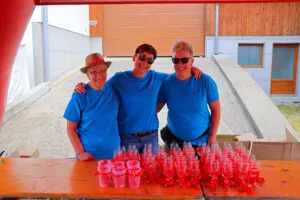 Trois femmes souriantes portant des t-shirts bleus se tiennent derrière une table avec de nombreux verres et gobelets rouges lors d'un événement en plein air.